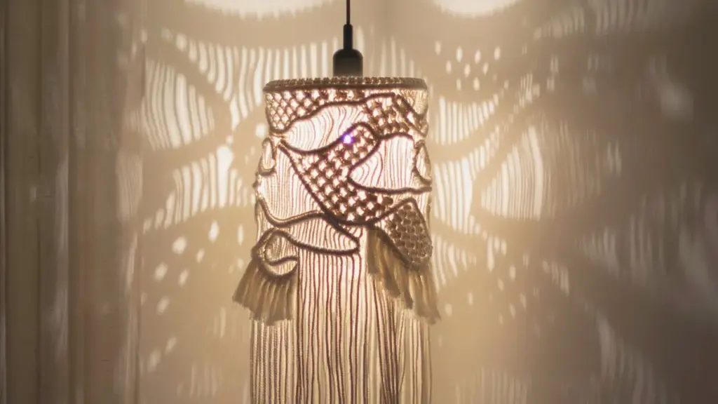 Lámpara de macramé encendida, hecha con nudos y tramas curvas, proyectando sombras decorativas sobre la pared.