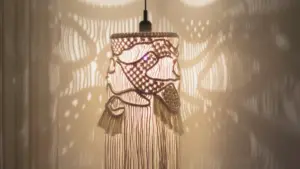 Lámpara de macramé encendida, hecha con nudos y tramas curvas, proyectando sombras decorativas sobre la pared.